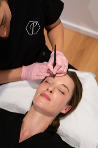 Woman lying with eyes closed receiving eyebrow treatment from a clinician wearing pink gloves