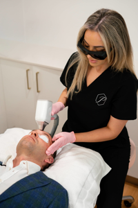 Clinician wearing black uniform and pink gloves performing laser skin treatment on man's face with eye protection